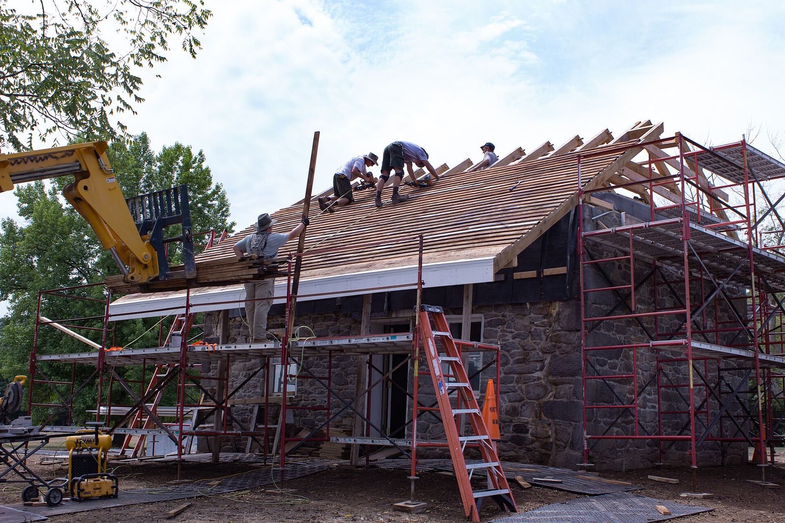 Roofing crew decking a steep roof on a stone building, a telehandler lifting the bundles